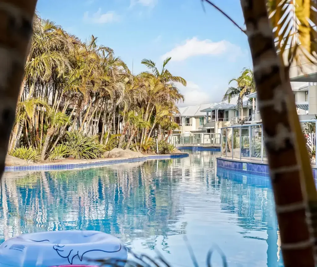 Resort style lagoon pool at a Port Stephens hotel surrounded by palm trees lush tropical gardens and modern apartment balconies. Calm blue water reflects the greenery and buildings creating a relaxed setting for a Port Stephens family holiday.
