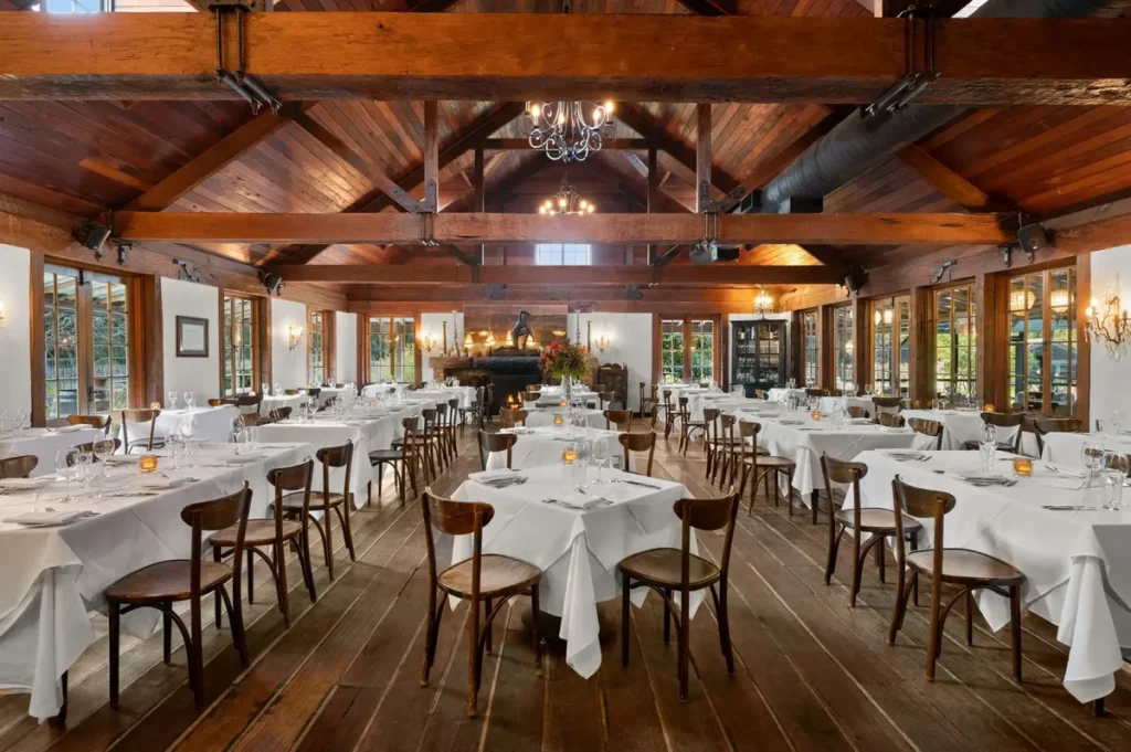 Elegant dining room at Circa 1876 featuring exposed timber beams polished wooden floors white tablecloths and a fireplace creating a warm heritage atmosphere. The refined restaurant offers a memorable dining experience during a Hunter Valley family holiday.