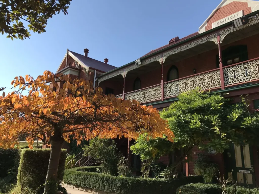Red brick Alzburg Resort building with an ornate lacework balcony and leafy gardens, framed by a small tree with golden autumn leaves. Signs reading "CONVENT" and "RECEPTION" are visible, and the heritage setting makes it a charming base for a Victorian High Country family holiday.