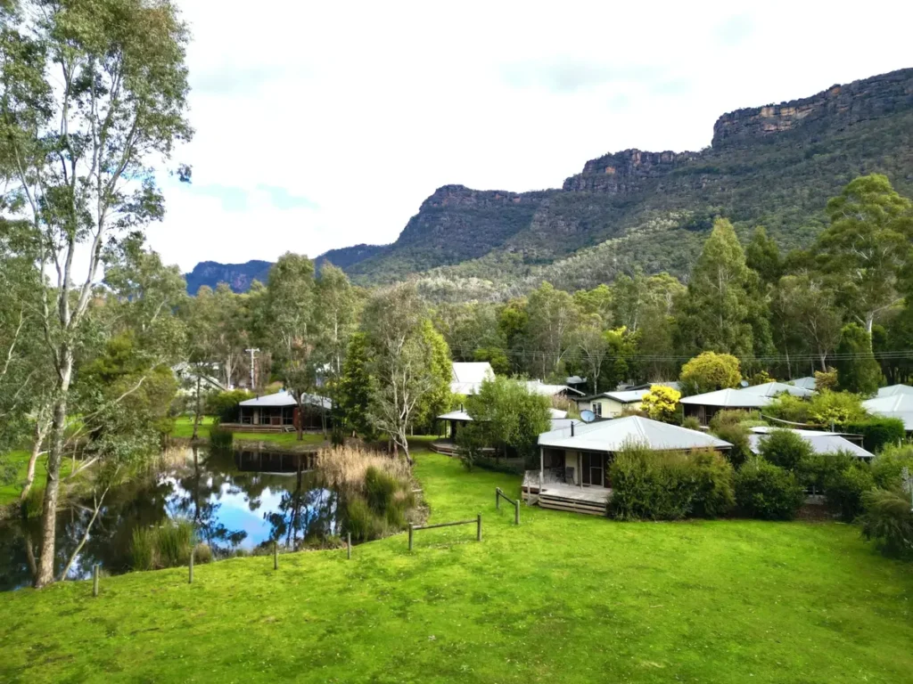 Grampians chalets surrounded by green lawns and native trees beside a reflective pond. Rugged mountain cliffs rise behind the accommodation and create a peaceful base for a Grampians family holiday.