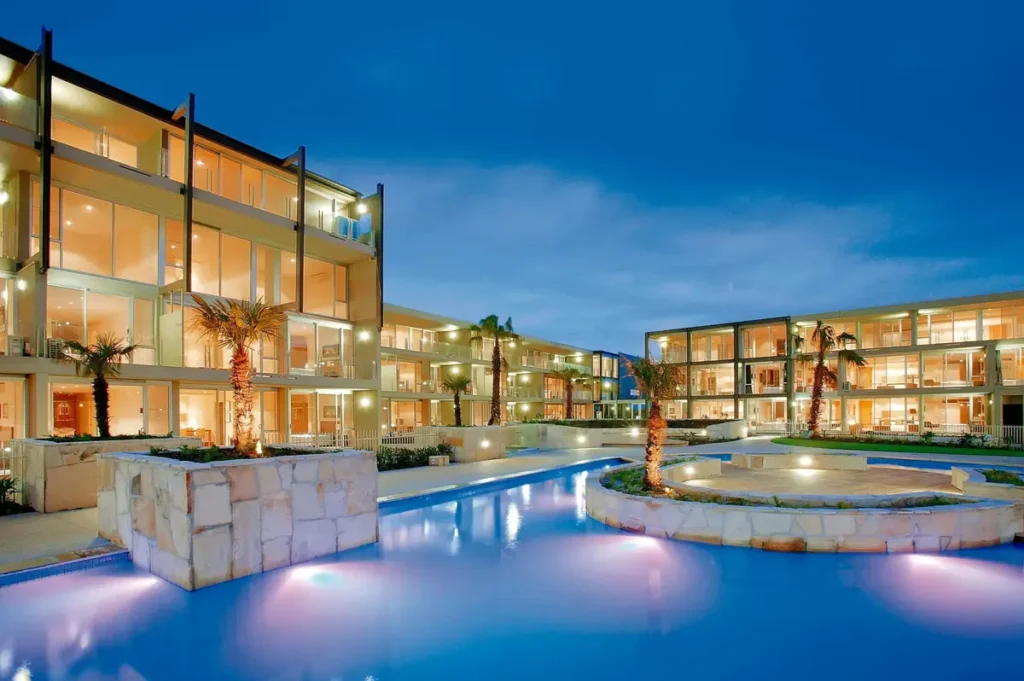 Wyndham Resort Torquay at dusk with illuminated apartments surrounding a curving blue swimming pool and palm trees. The spacious resort setting offers a relaxing base for a Great Ocean Road family holiday.