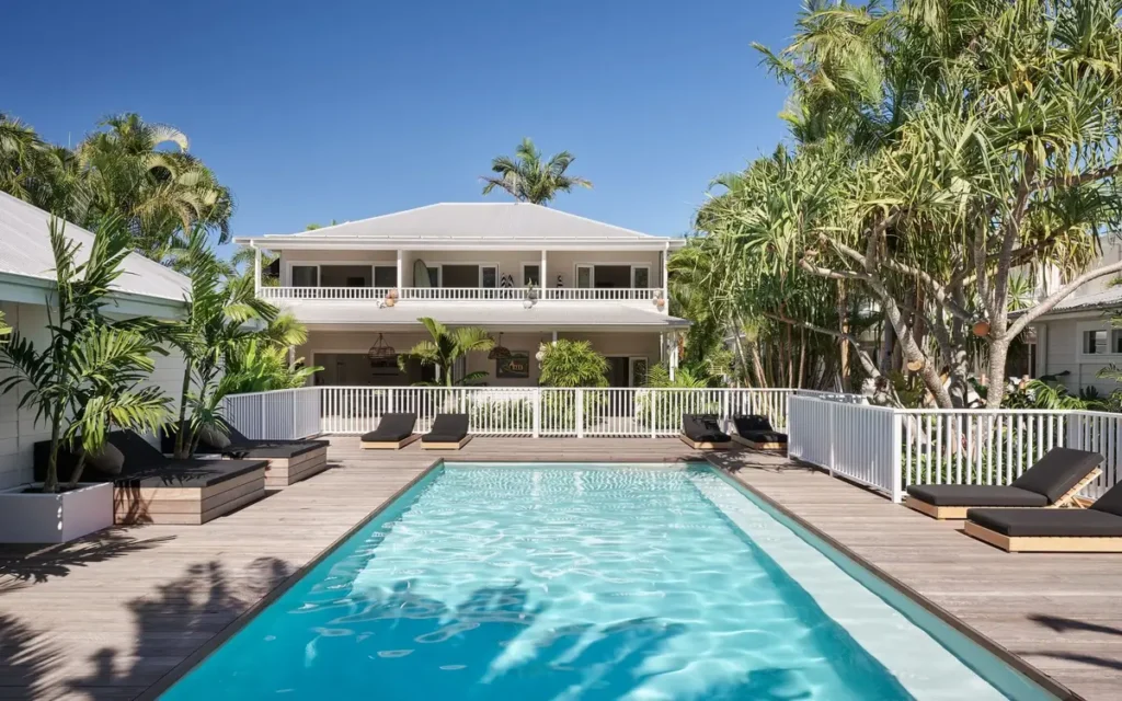 The Atlantic Byron has a long turquoise pool with sun loungers, tropical plants, and a white two story guesthouse behind it. This relaxed Byron Bay accommodation makes a calm final stop on a Gold Coast to Byron Bay family holiday.