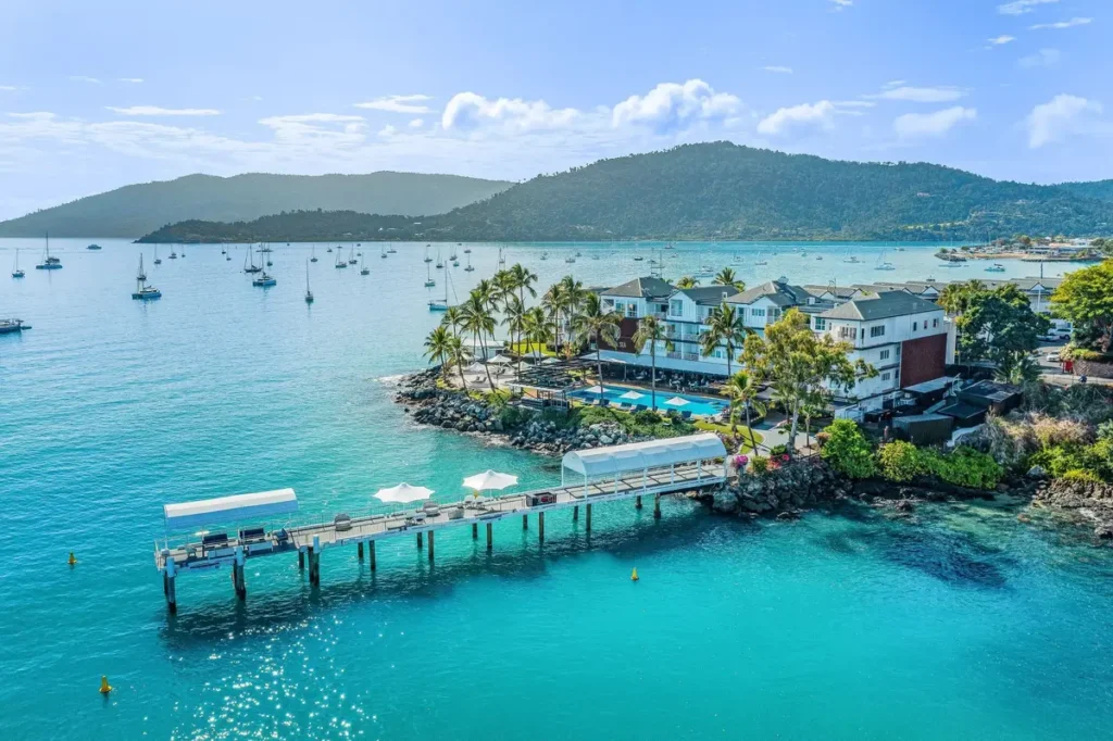 Turquoise water surrounds Coral Sea Marina with sailboats anchored near a waterfront resort and green hills in the background. It shows a calm coastal base for a Whitsunday Coast family holiday.