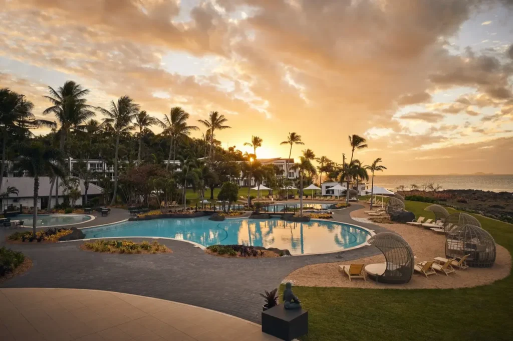 Palm trees and resort pools glow at sunset beside the ocean on Daydream Island. The peaceful tropical setting suits a relaxed Whitsunday Coast family holiday.