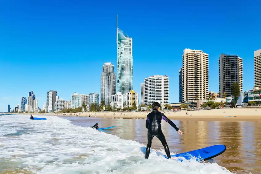 A surfer in a black wetsuit stands on a blue board in the shallow waves at the Gold Coast, with tall beachfront skyscrapers behind them under a clear blue sky. This beach scene highlights the surfing culture that makes a Gold Coast to Byron Bay family holiday so appealing.