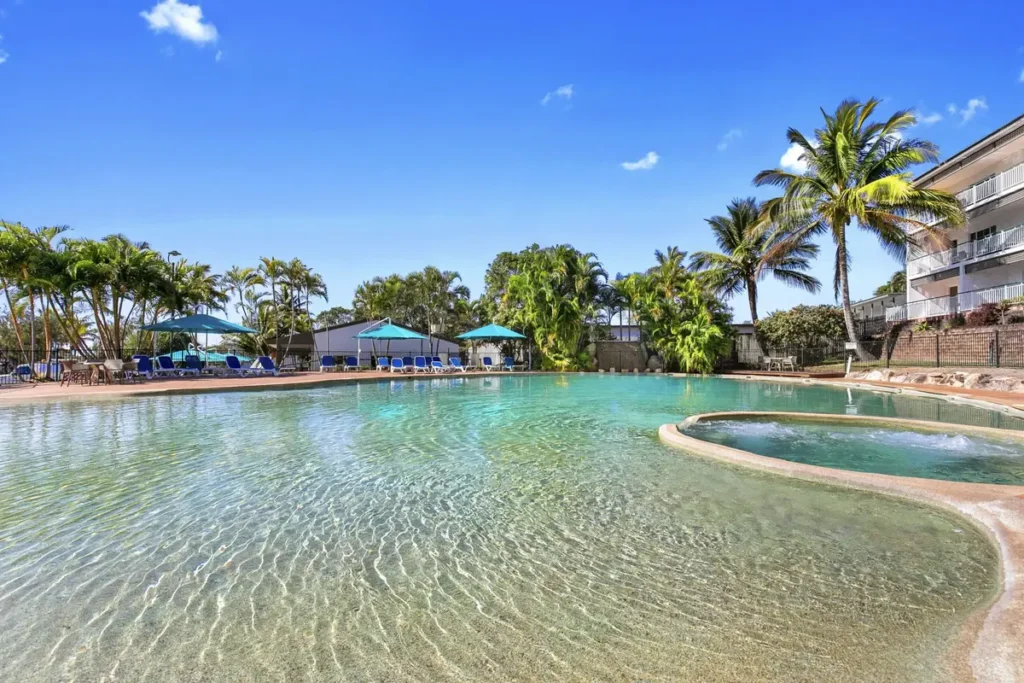 Kโgari Beach Resort has a large lagoon style pool with a spa, blue umbrellas, palm trees, and resort buildings nearby. The family friendly pool area offers a relaxed place to stay during a Fraser Island family holiday.