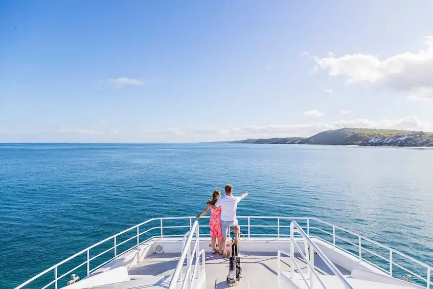 Two people stand at the front of a boat looking across calm blue water toward the coastline of Kโgari. The boat tour offers a scenic way to explore the island during a Fraser Island family holiday.