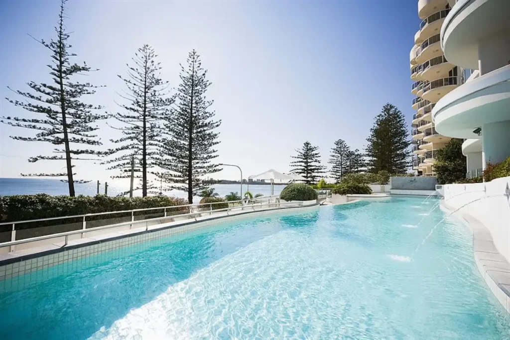 Mantra Sirocco has a bright lagoon style pool beside curved apartment balconies, tall pine trees, and ocean views. The beachfront resort setting offers an easy Sunshine Coast family holiday base.