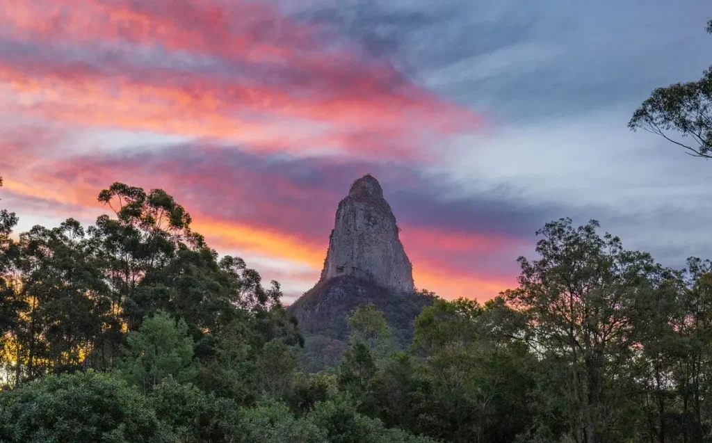 Mount Coonowrin rises above dark green bushland under a dramatic sunset sky streaked with pink, orange, and blue. This striking Glass House Mountains view adds a memorable scenic stop to a Sunshine Coast Hinterland family holiday.