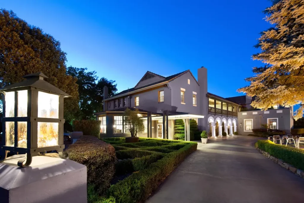 Peppers Mineral Springs at dusk with warmly lit windows. Manicured hedges and an illuminated driveway create a welcoming setting for a Macedon Ranges family holiday.