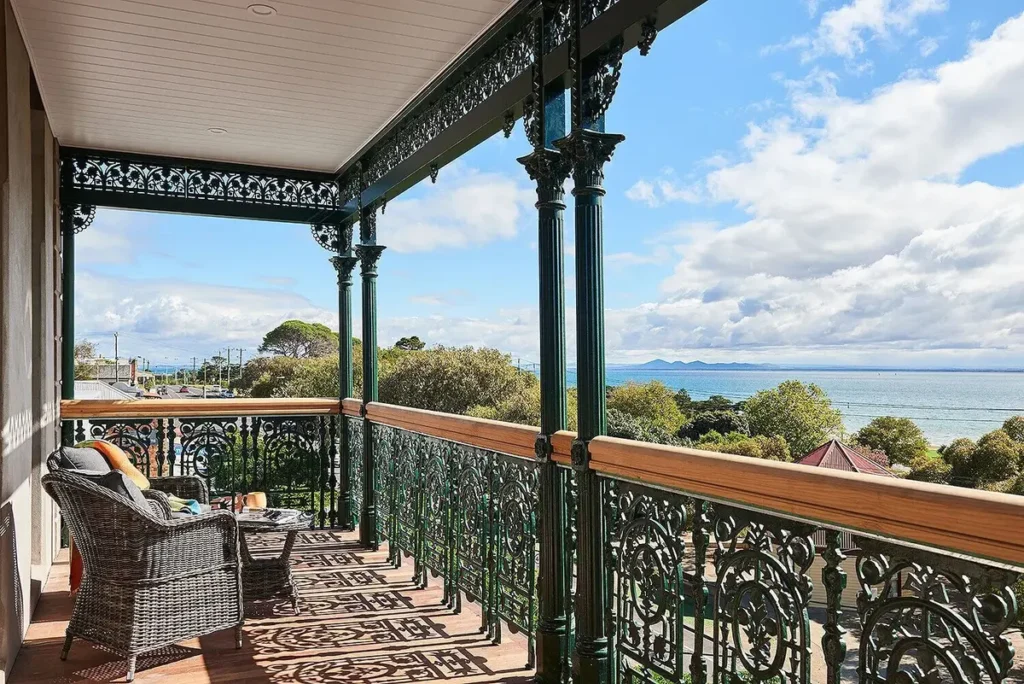 Ornate balcony at the Portarlington Grand Hotel with wicker chairs overlooking treetops and the blue waters of Port Phillip Bay. The coastal view offers a relaxing setting for a Bellarine Peninsula family holiday.