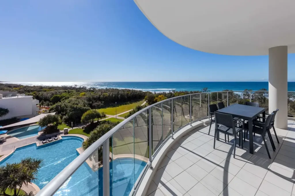 A curved balcony at Ramada overlooks resort pools, green gardens, and the ocean beyond the dunes. The spacious coastal view offers a relaxed Sunshine Coast family holiday base.