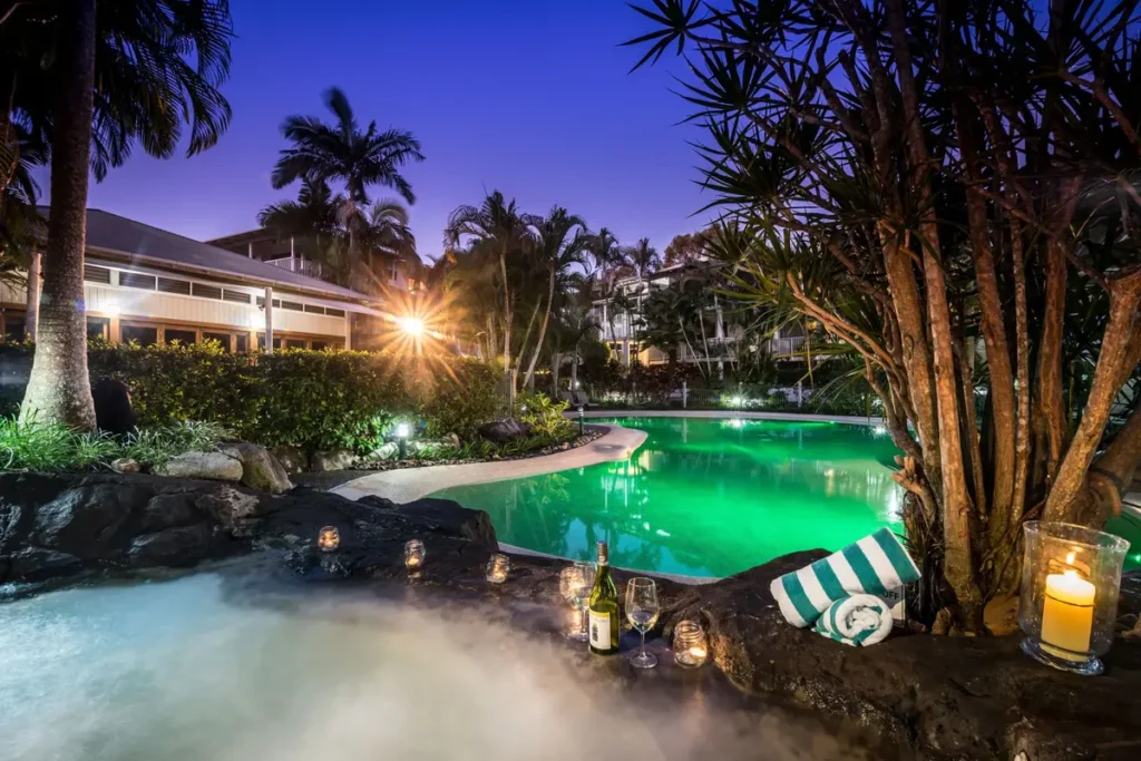 South Pacific Resort has a glowing lagoon pool and spa surrounded by palm trees, candles, and tropical gardens at dusk. The relaxed resort setting offers a peaceful stay during a Great Beach Drive family holiday.