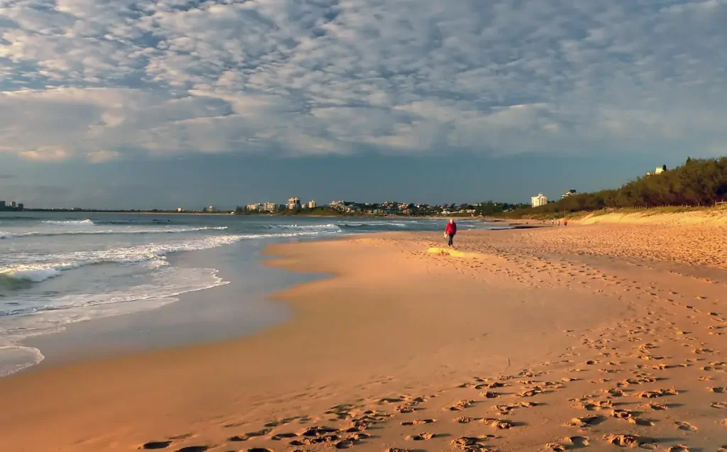 Golden light falls across a wide Sunshine Coast beach with gentle waves, footprints in the sand, and a few people walking near the water. The peaceful shoreline captures an easy beach outing during a Sunshine Coast family holiday.