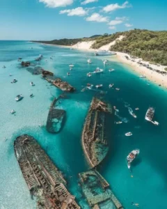 Shipwrecks at Moreton Island in clear turquoise water near a white sand beach. Small boats and swimmers surround the wrecks along the sunny coastline.