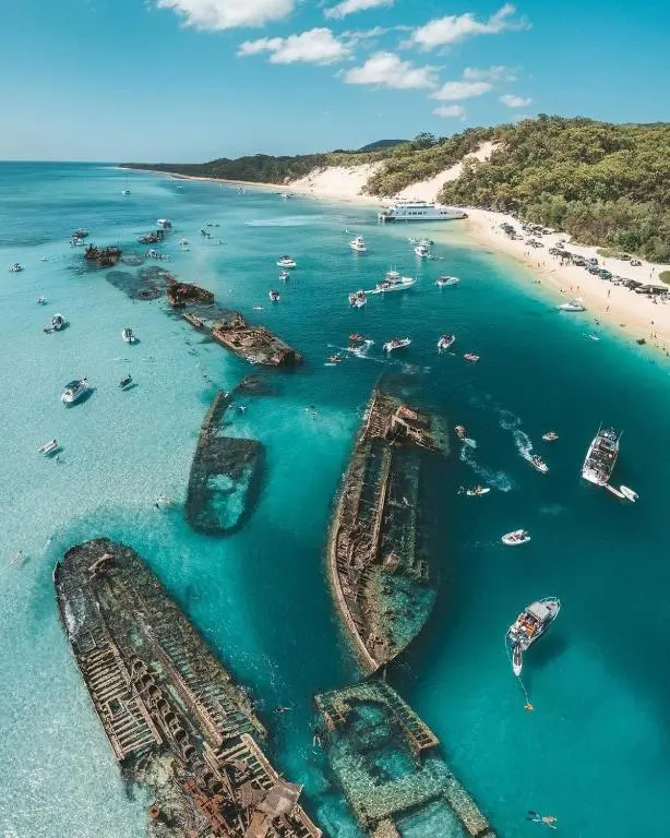 Shipwrecks at Moreton Island in clear turquoise water near a white sand beach. Small boats and swimmers surround the wrecks along the sunny coastline.