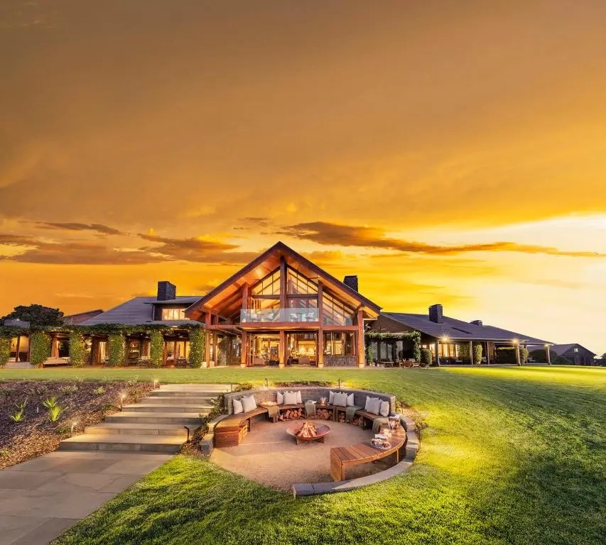 Spicers Peak Lodge glows at sunset with a large glass front, wide green lawn, and circular outdoor fire pit. The luxury country retreat adds a scenic stay option to a Southern Downs and Granite Belt family holiday.