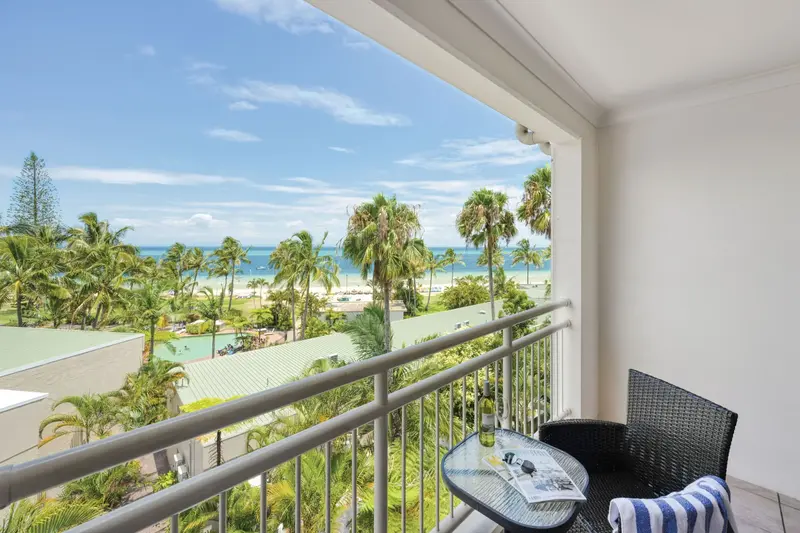 Balcony at Tangalooma Island Resort with a chair and table overlooking palm trees. The beach and bright blue ocean are visible beyond the resort grounds.