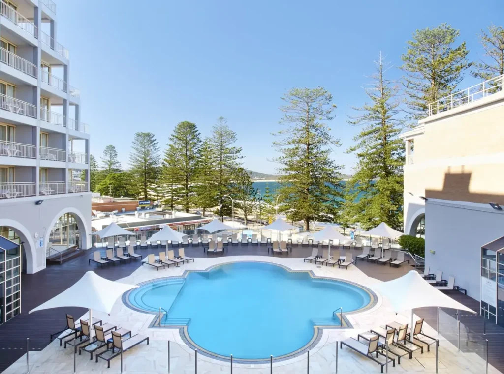 Crowne Plaza Terrigal Pacific featuring a curved outdoor swimming pool surrounded by sun loungers white shade sails and modern hotel buildings overlooking the ocean. The beachfront location and resort style amenities make it a popular choice for a Central Coast family holiday.