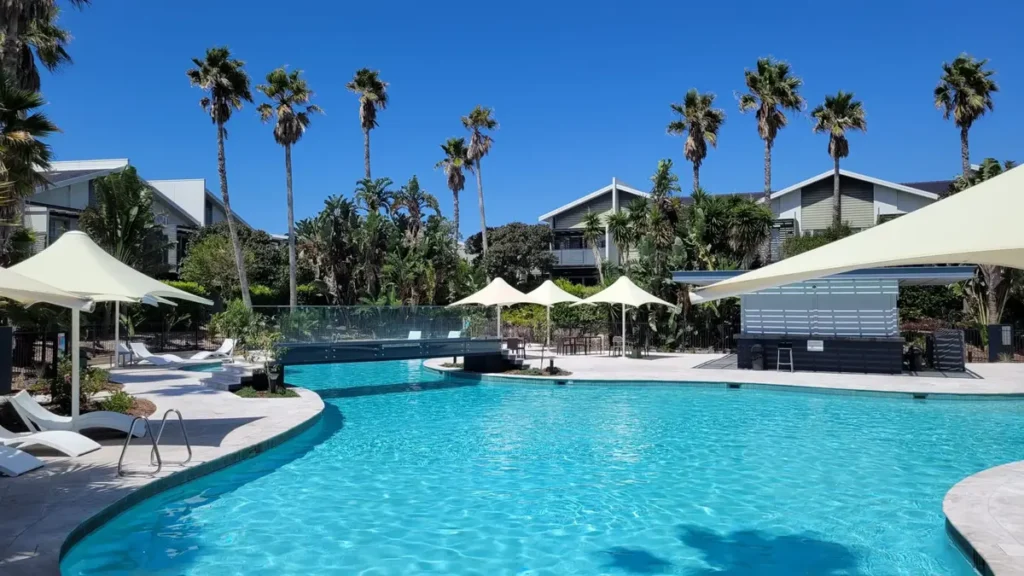 Pullman Magenta Shores Resort on the Central Coast featuring a large lagoon style swimming pool surrounded by palm trees shaded loungers and modern apartment buildings. The relaxed resort setting makes it an ideal base for a Central Coast family holiday.
