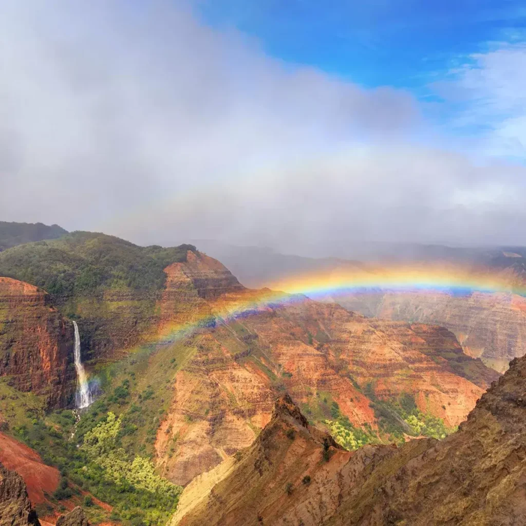 A vibrant rainbow arcs across the sky above Waimea Canyon, also known as the "Grand Canyon of the Pacific," in Hawaii. The deep red and green canyon walls contrast with the cascading waterfall in the distance. A misty sky blends into the blue horizon, creating a surreal and magical view.