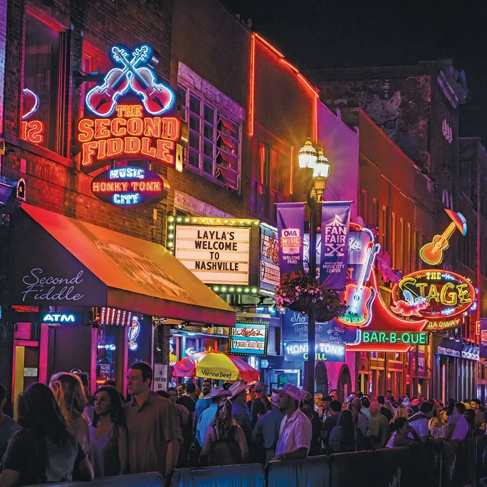 A lively night scene on Broadway Street in Nashville, Tennessee, with neon lights illuminating the street. Signs for famous honky-tonk bars like "The Second Fiddle" and "The Stage" glow against the dark sky. A crowd of people fills the street, enjoying the bustling nightlife of Music City.