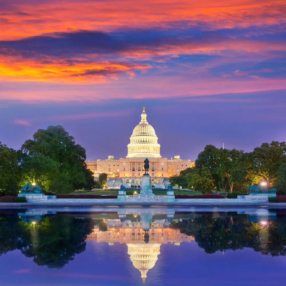 The U.S. Capitol building in Washington, D.C., illuminated against a vibrant sunset sky with shades of orange, pink, and purple. The reflection of the historic landmark shimmers in the water of the Capitol Reflecting Pool, adding symmetry to the scene. Lush green trees and statues frame the foreground, enhancing the grandeur of the iconic structure.