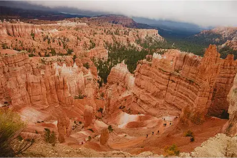 A breathtaking view of Bryce Canyon National Park in Utah, showcasing towering red rock formations known as hoodoos. A winding trail cuts through the canyon, with tiny hikers making their way along the path. A misty sky looms above, adding a dramatic contrast to the earthy landscape.