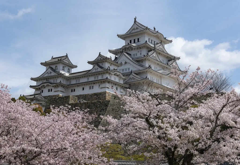 A white Japanese castle with layered tiled roofs stands on a stone base, surrounded by pale pink cherry blossom trees in full bloom. The scene captures Cherry Blossom Season in Japan with spring flowers framing the historic architecture under a clear blue sky.