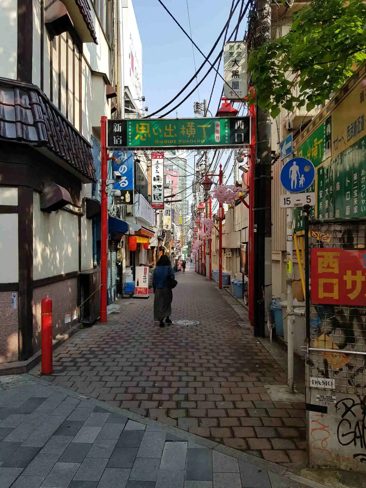 A narrow pedestrian alleyway in Tokyo’s Shinjuku district features the entrance to Omoide Yokocho, marked by a green sign with yellow and white Japanese characters and the English translation below. The street is lined with small restaurants and izakayas, red lanterns, and overhead wires, with a lone woman walking into the alley.