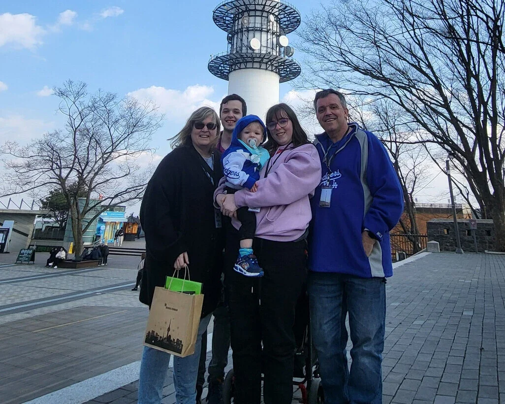 Three generations of a family pose together during a multi generational travel outing in front of a tall observation tower. Two older adults, two younger adults, and a baby dressed in blue stand close together smiling, with bare trees and an open plaza visible behind them.