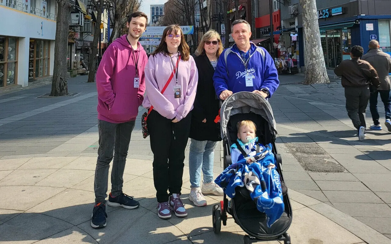 Three generations of a family stand together on a sunny city street during a multi generational travel outing. Two younger adults, an older couple, and a baby in a stroller smile at the camera with shops and bare trees lining the background.