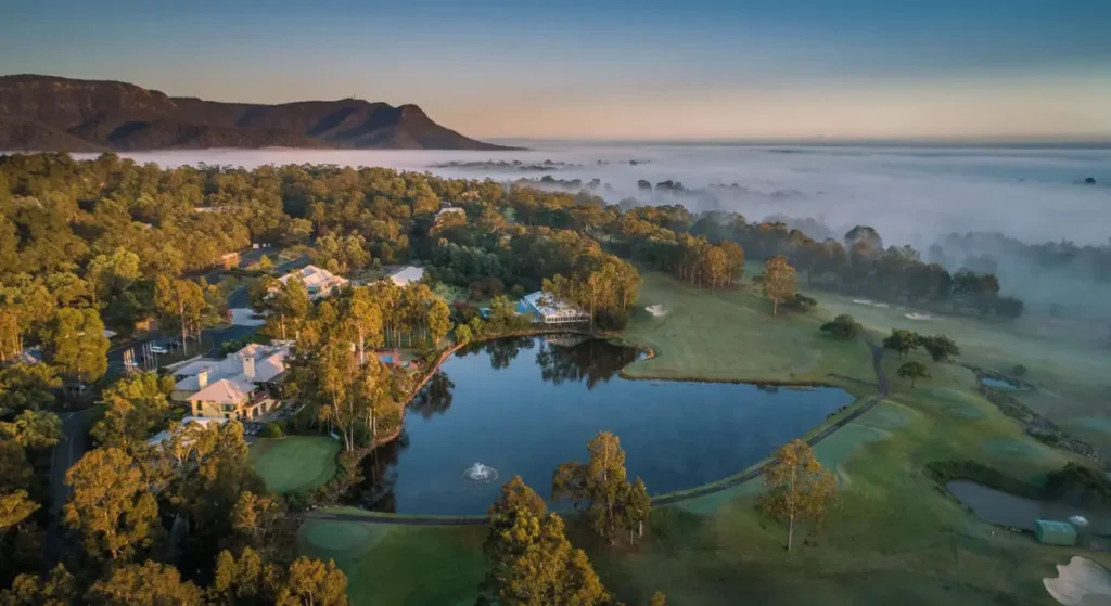 Aerial view of Oaks Cypress Lakes Resort in the Hunter Valley surrounded by rolling golf course greens a large lake and misty countryside at sunrise. The scenic vineyard setting offers spacious accommodation for a Hunter Valley family holiday.