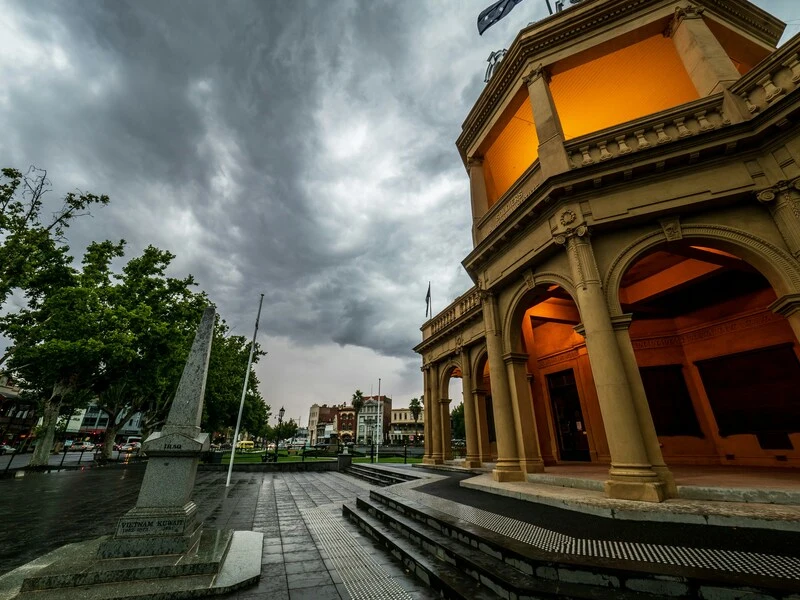 Historic civic building in Bendigo illuminated with warm orange light under dark storm clouds. A stone war memorial stands in the foreground on a wet plaza, creating a dramatic contrast between architecture and weather. This image sets the mood for exploring the city and supports content about the Best Way to See Victoria on a Short Trip.