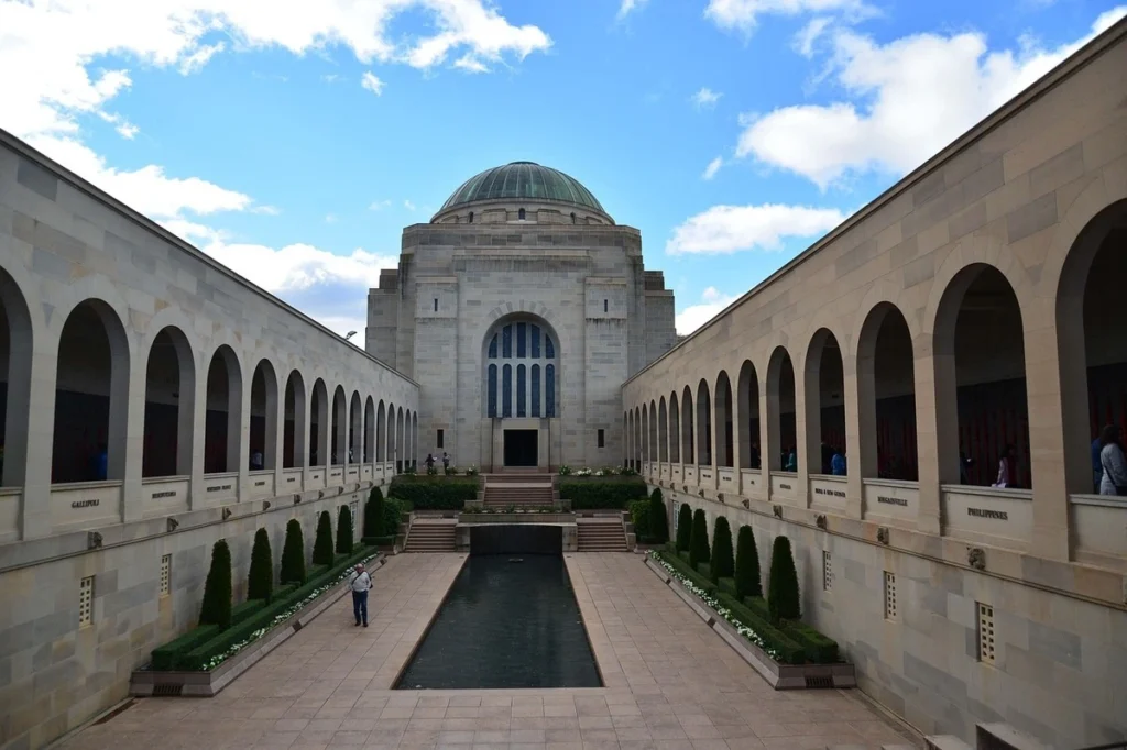 View of the Australian War Memorial in Canberra featuring a long reflective pool symmetrical stone archways and a domed building at the end. Visitors walk through the courtyard during a Canberra family holiday exploring national history and landmarks.