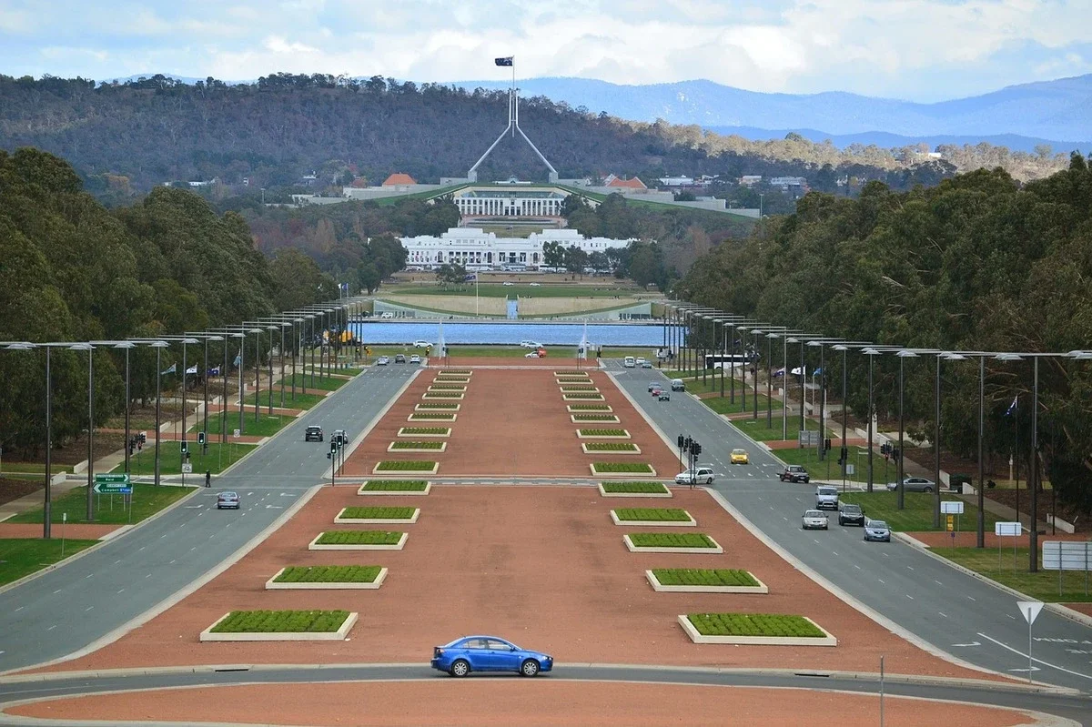 Wide view of Parliament House in Canberra seen from across a landscaped boulevard with symmetrical garden beds and light traffic. The iconic building sits beneath rolling hills and open sky during a Canberra family holiday visit.