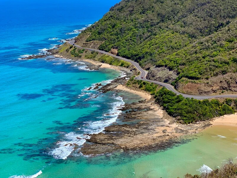 Winding coastal road hugging a steep green hillside beside bright turquoise ocean water. Waves break against rocky shorelines and small beaches along the Great Ocean Road. This image highlights a scenic drive that is essential to the Best Way to See Victoria on a Short Trip.
