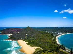 Aerial view of a coastal headland in Port Stephens with golden sandy beaches turquoise ocean and lush green hills stretching toward a seaside town. The sweeping shoreline and clear blue water create a scenic backdrop for a Port Stephens family holiday.