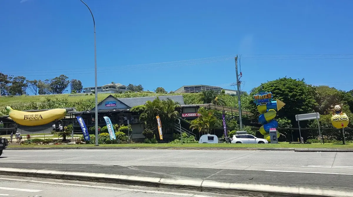Roadside view of The Big Banana attraction with a large banana sculpture colorful Water Park sign and laser tag entrance along a sunny street. Cars and palm trees sit in front of the entertainment complex on a grassy hillside.