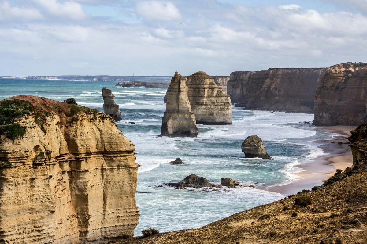 Limestone sea stacks rising from the ocean along the Twelve Apostles coastline in Victoria. Waves crash against the cliffs and sandy shore under a partly cloudy sky. This iconic landscape represents a must see stop when planning the Best Way to See Victoria on a Short Trip.