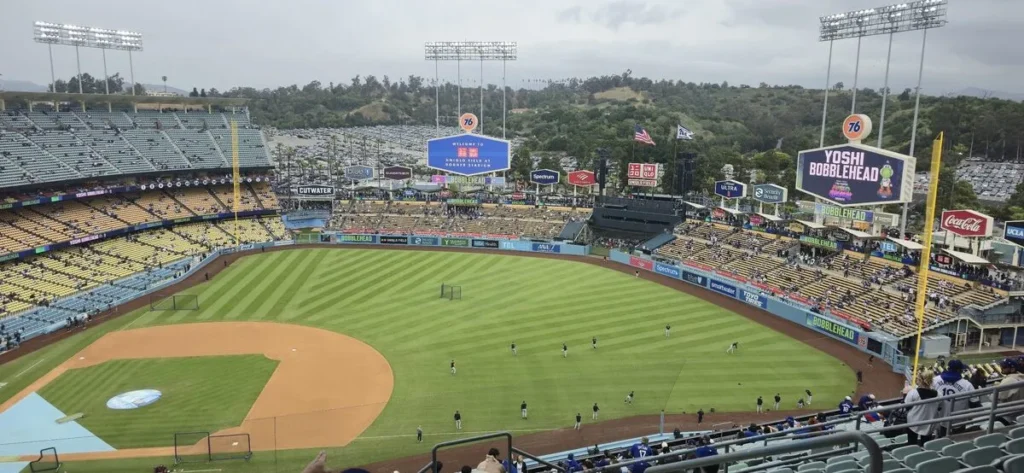 Wide view of Dodger Stadium in Los Angeles with players warming up on the field and fans seated in the stands. Scoreboards and surrounding hills frame the iconic baseball stadium setting.