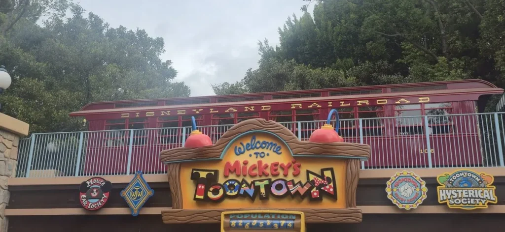 Welcome to Mickey’s Toontown sign at Disneyland with colorful cartoon lettering and a red Disneyland Railroad train in the background. Playful themed signage and character logos create a lively family friendly theme park atmosphere.