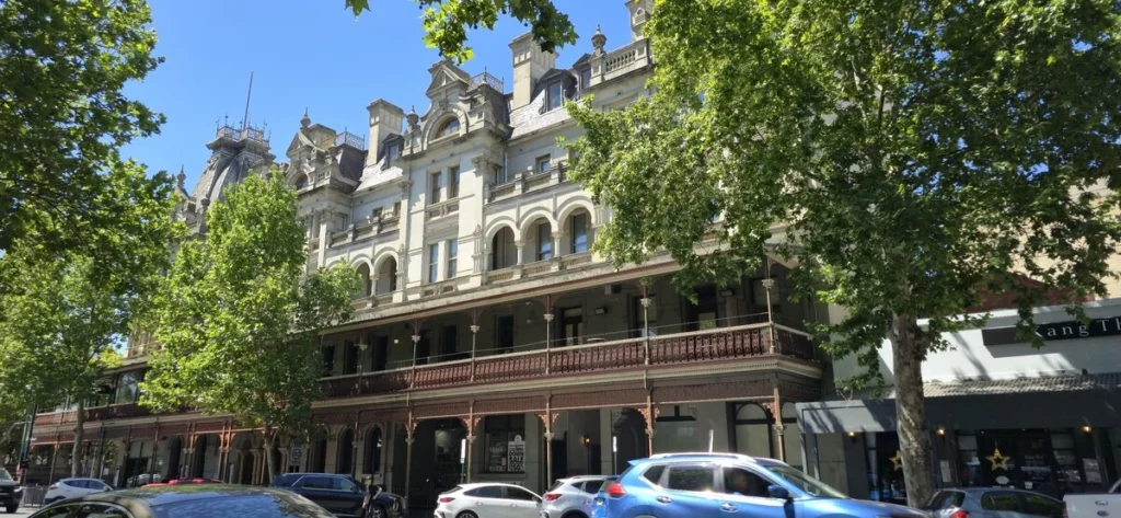 Historic Shamrock Hotel in Bendigo with ornate cream stonework and a long iron lace balcony shaded by leafy trees. The grand heritage building is a distinctive city landmark to see on a Bendigo family holiday.