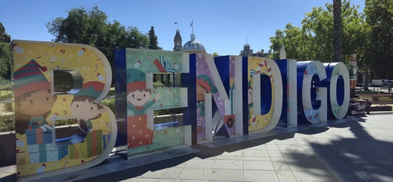 Large colourful letters spelling "BENDIGO" decorated with festive characters and gifts in a sunny city park. The playful landmark marks a cheerful stop on a Bendigo family holiday.